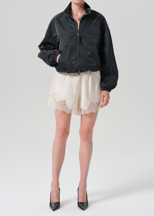 A model wearing a black zip-up bomber jacket, cream lace-trimmed shorts, and black pointed-toe heels, photographed against a grey background.