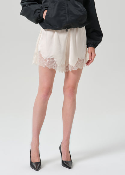 A model is shown from the waist down wearing a black jacket, cream satin shorts with a lace hem, and black pointed-toe heels, set against a plain grey background.