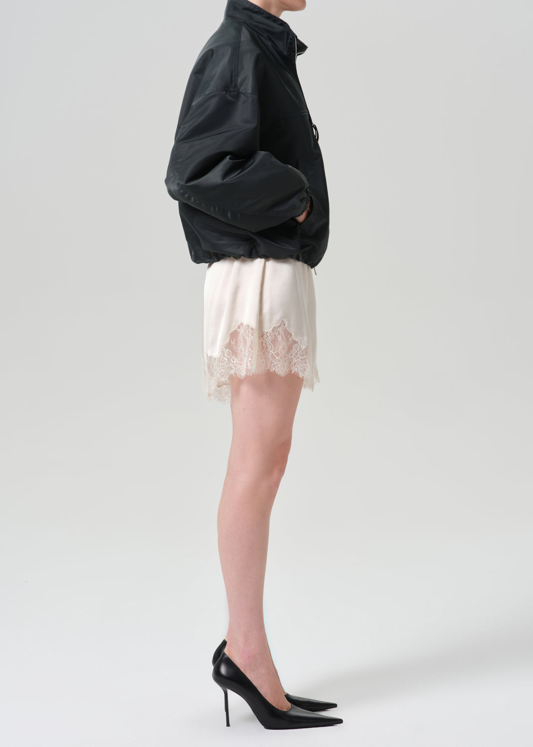 A model shown from the side wearing a black jacket, a cream satin mini skirt with a lace hem, and black pointed-toe stiletto heels against a grey background.