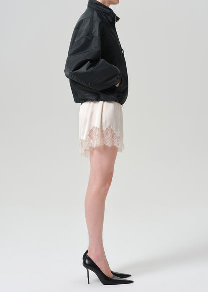 A model shown from the side wearing a black jacket, a cream satin mini skirt with a lace hem, and black pointed-toe stiletto heels against a grey background.