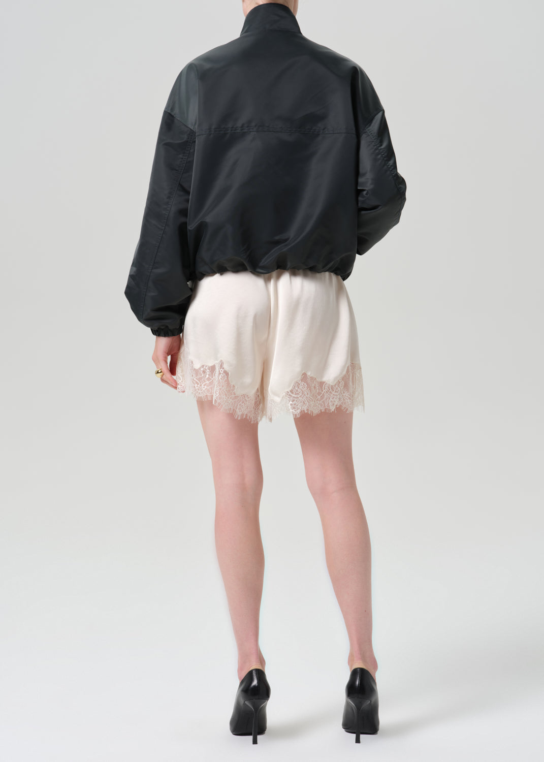 A model shown from the back wearing a black bomber jacket, cream lace-trimmed shorts, and black high heels against a grey background.
