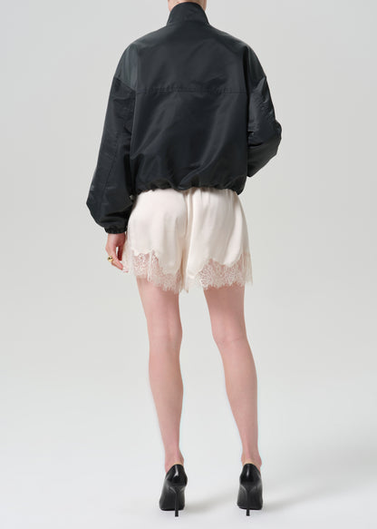 A model shown from the back wearing a black bomber jacket, cream lace-trimmed shorts, and black high heels against a grey background.