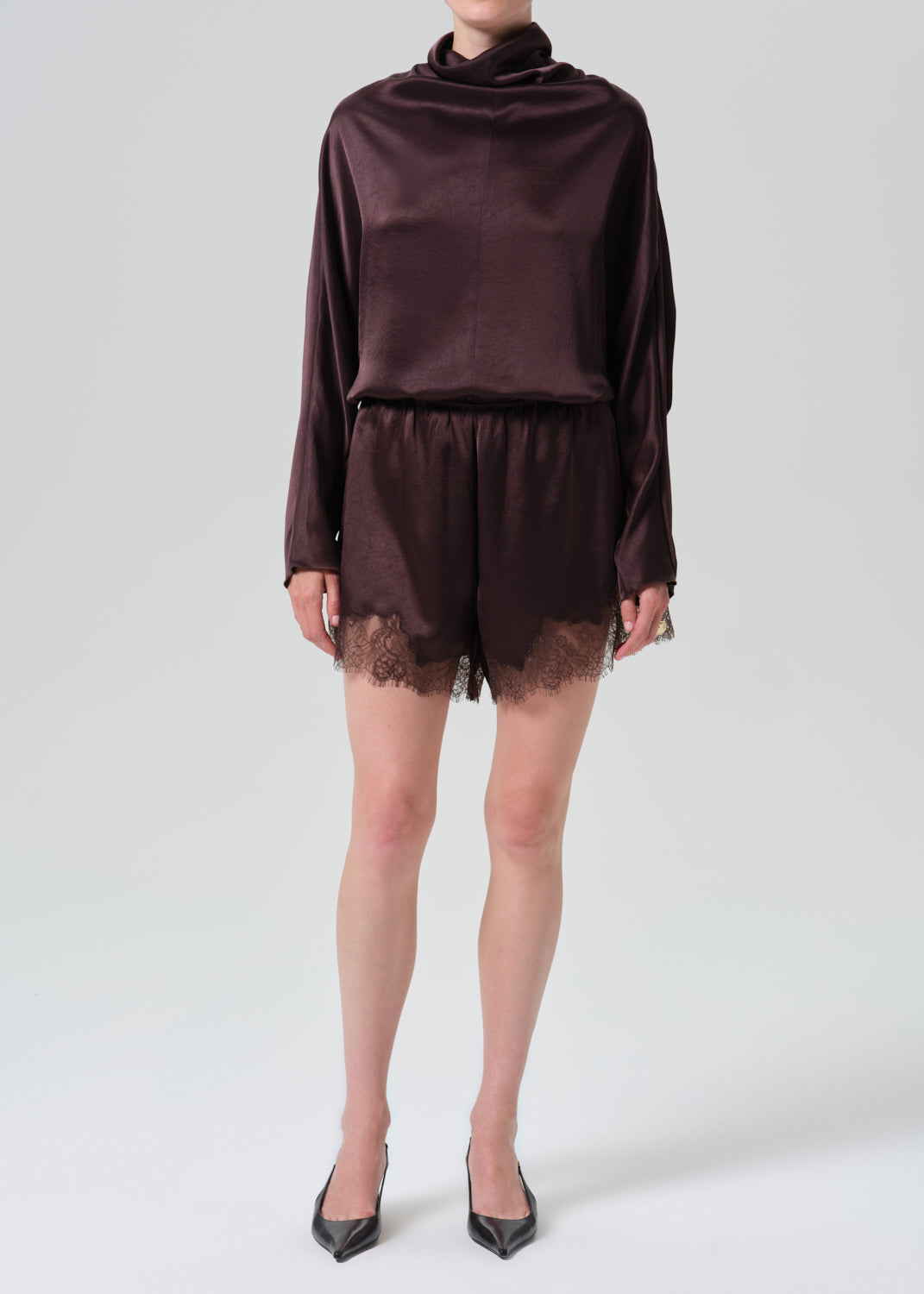 A model wearing a dark plum satin long-sleeve top and matching satin shorts with lace trim, paired with black pointed-toe heels, against a grey background.