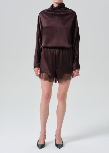 A model wearing a dark plum satin long-sleeve top and matching satin shorts with lace trim, paired with black pointed-toe heels, against a grey background.