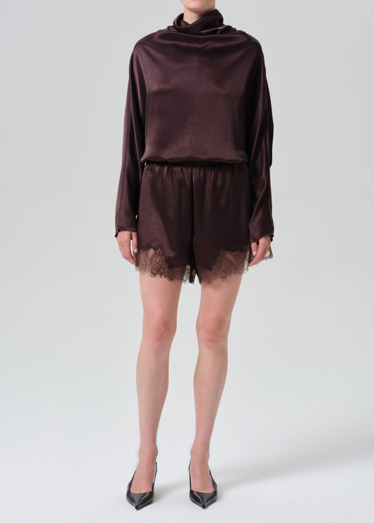 A model wearing a dark plum satin long-sleeve top and matching satin shorts with lace trim, paired with black pointed-toe heels, against a grey background.