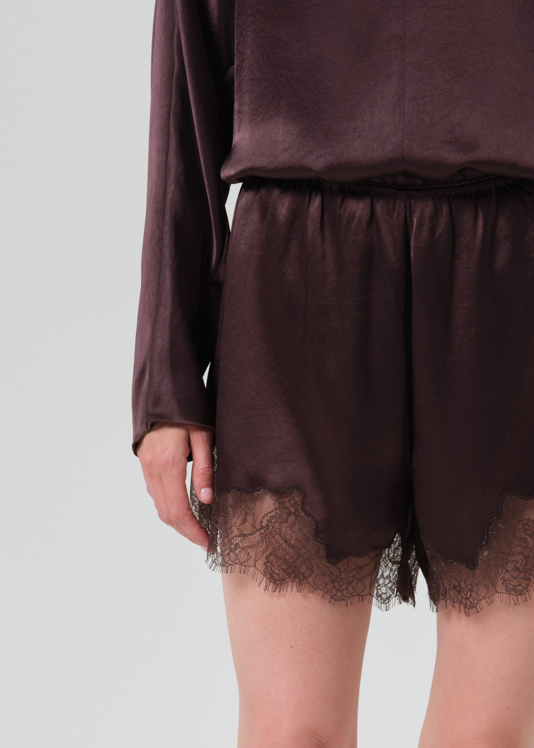 A close-up of a person wearing a dark plum satin-like two-piece outfit with lace-trimmed shorts and a matching long-sleeve top against a grey background.