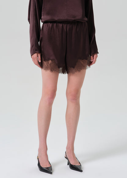 A model wearing a dark brown satin-like two-piece set consisting of a long-sleeve top and matching shorts with a lace hem, paired with black pointed-toe heels against a grey background.