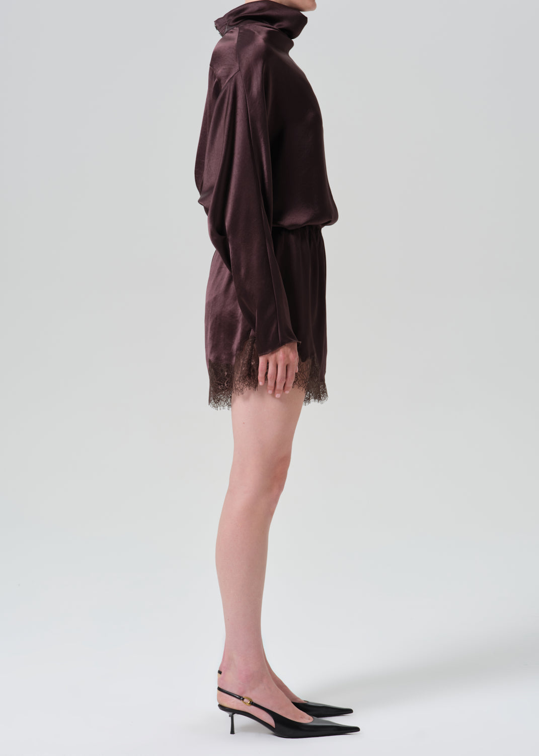 A side-profile studio shot of a model wearing a dark plum purple satin mini dress with a turtleneck, long draped sleeves, and a lace-trimmed hem, paired with black slingback heels.