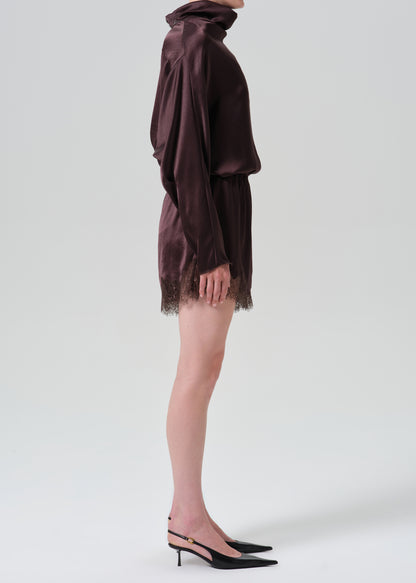 A side-profile studio shot of a model wearing a dark plum purple satin mini dress with a turtleneck, long draped sleeves, and a lace-trimmed hem, paired with black slingback heels.