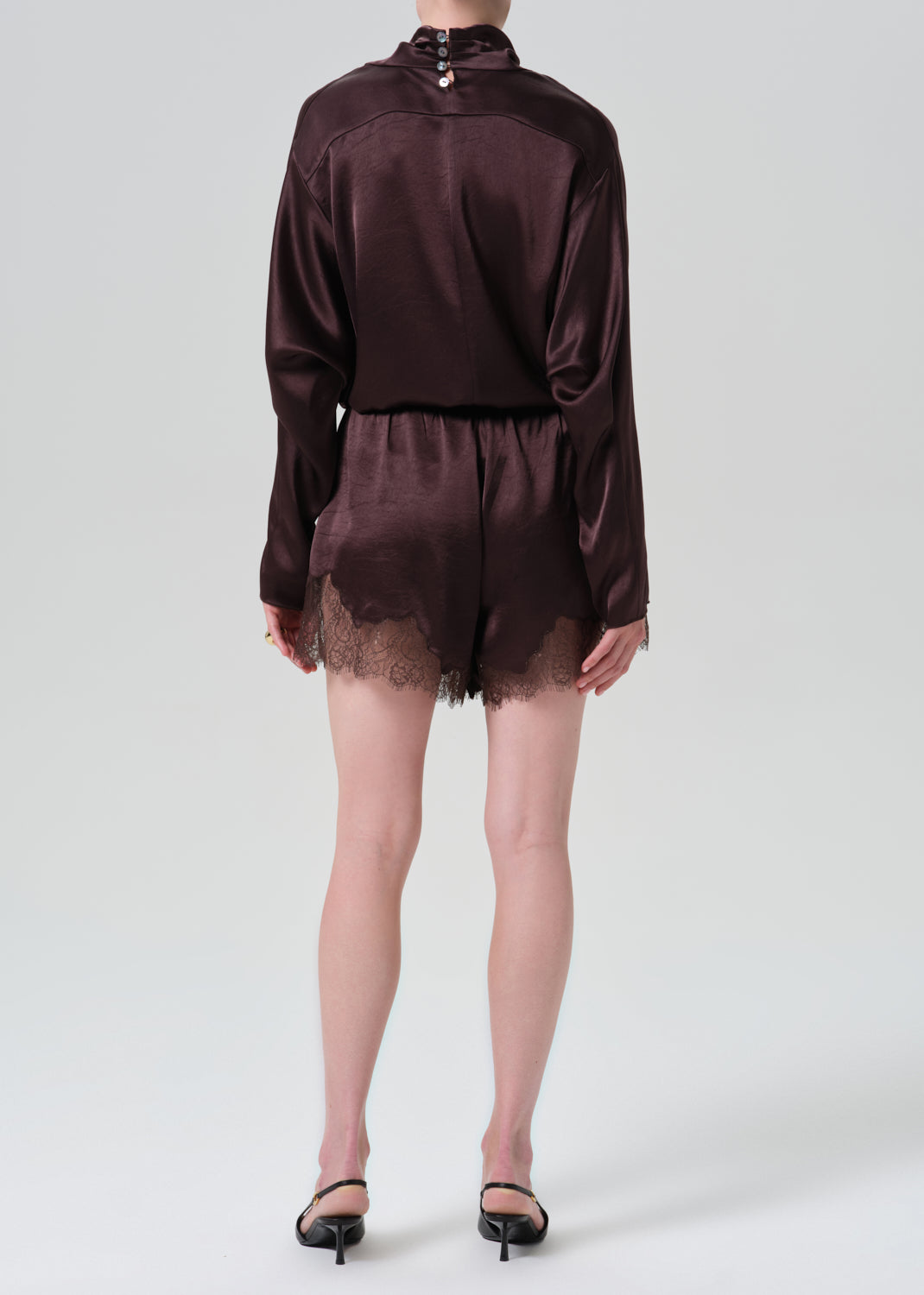 A model shown from the back wearing a dark brown satin long-sleeve romper with lace trim at the hem, paired with black pointed-toe heels, against a grey background.