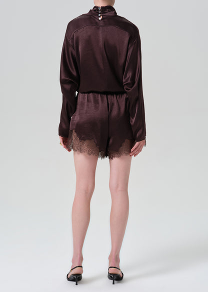 A model shown from the back wearing a dark brown satin long-sleeve romper with lace trim at the hem, paired with black pointed-toe heels, against a grey background.