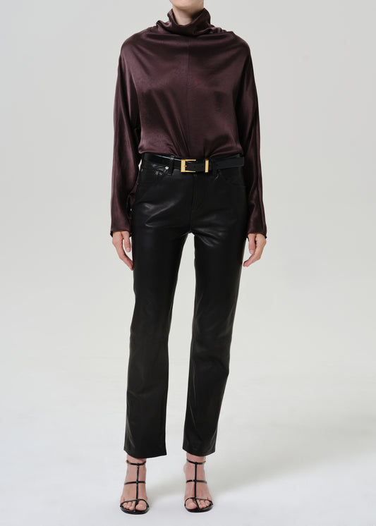 Woman wearing black leather pants with a dark brown satin draped neck top on a grey background.