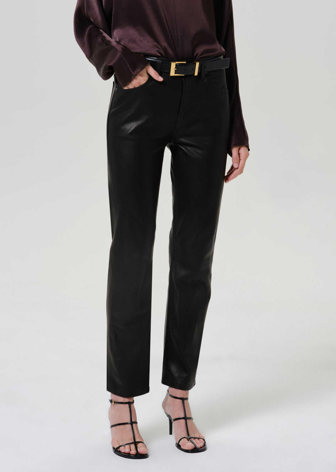 Woman wearing black leather pants with a dark brown satin draped neck top on a grey background.