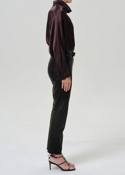Woman wearing black leather pants with a dark brown satin draped neck top viewed from the side on a grey background.