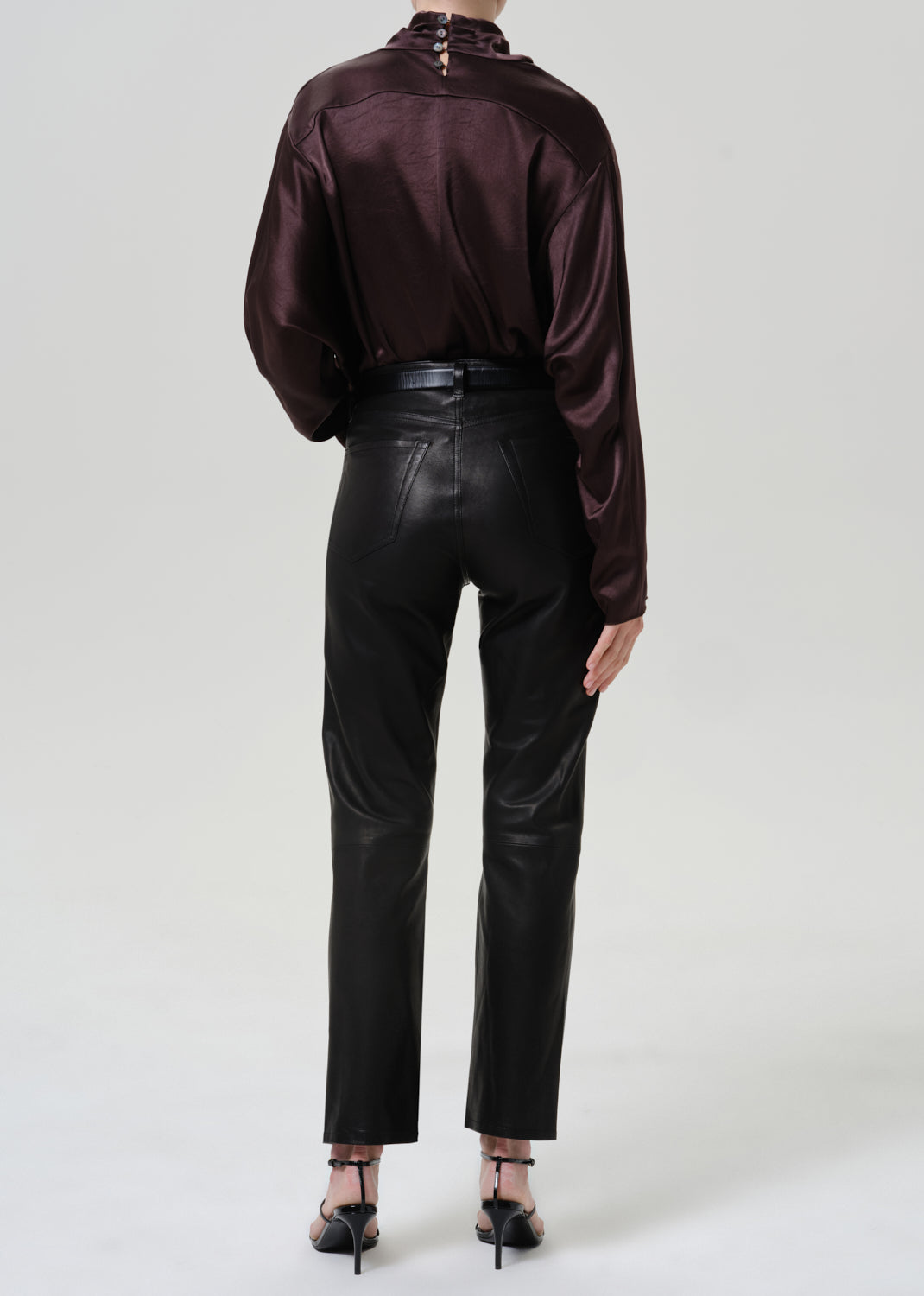 Woman wearing black leather pants with a dark brown satin draped neck top viewed from the back on a grey background.