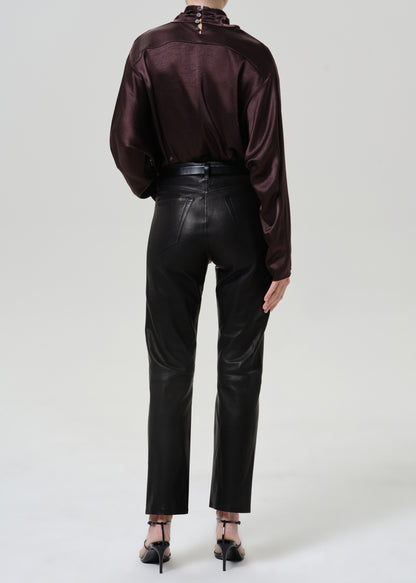 Woman wearing black leather pants with a dark brown satin draped neck top viewed from the back on a grey background.