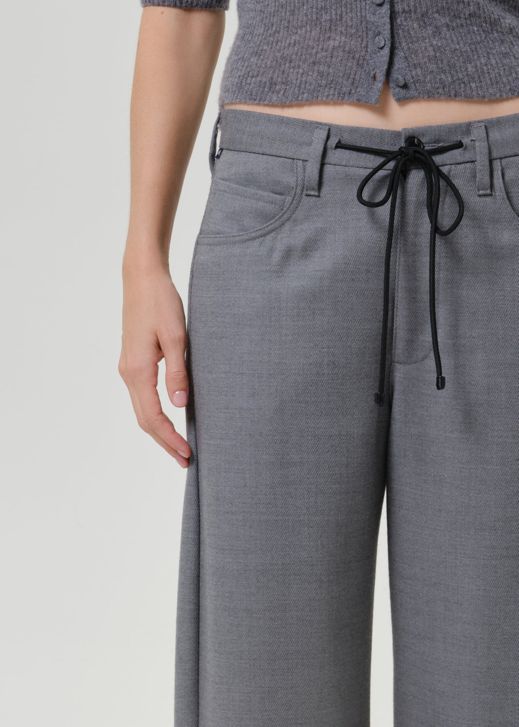 Close-up of a person wearing gray wide-leg trousers with a black drawstring waistband, paired with a gray cropped top, against a light gray studio background.