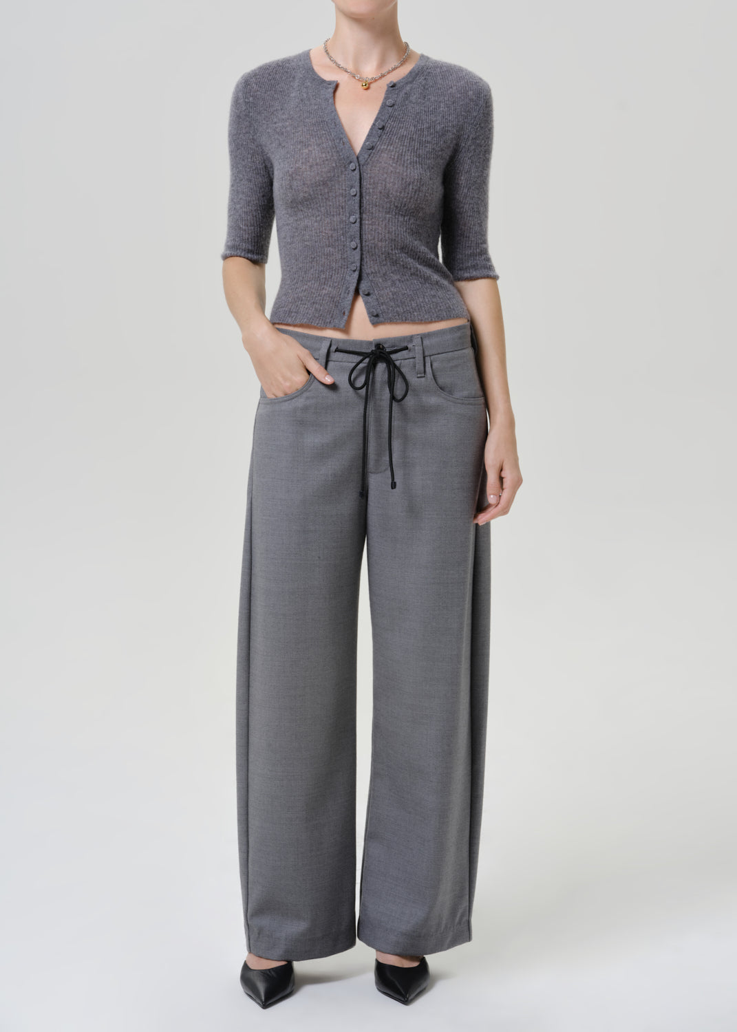 A model wearing a gray three-quarter sleeve button-up cardigan and matching gray wide-leg trousers with a drawstring waist, paired with black pointed-toe heels, against a light gray studio background.
