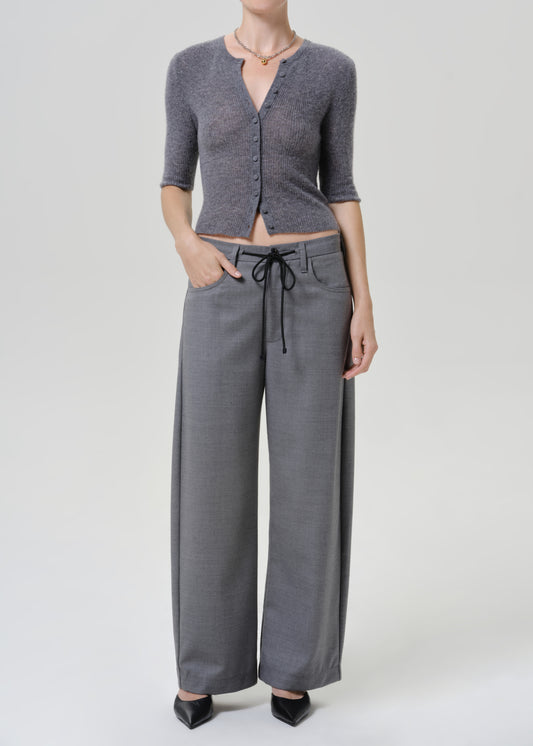 A model wearing a gray three-quarter sleeve button-up cardigan and matching gray wide-leg trousers with a drawstring waist, paired with black pointed-toe heels, against a light gray studio background.