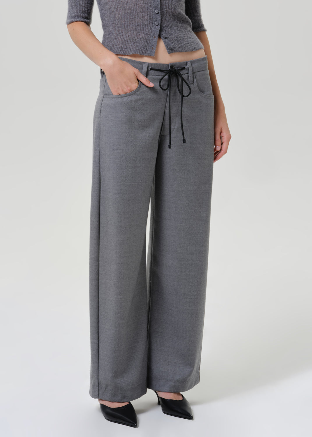 A model wearing a pair of wide-leg gray trousers with a black drawstring waist, paired with a gray cropped top and black pointed shoes, against a light gray studio background.