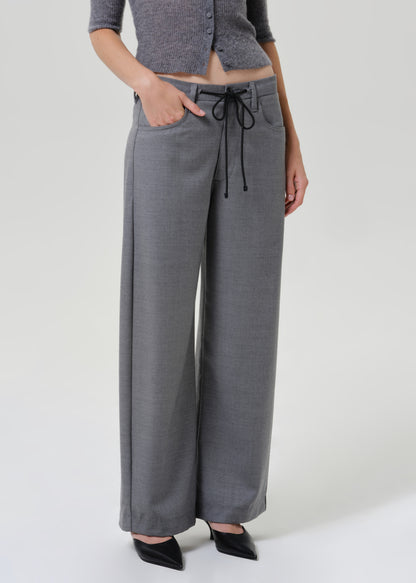 A model wearing a pair of wide-leg gray trousers with a black drawstring waist, paired with a gray cropped top and black pointed shoes, against a light gray studio background.
