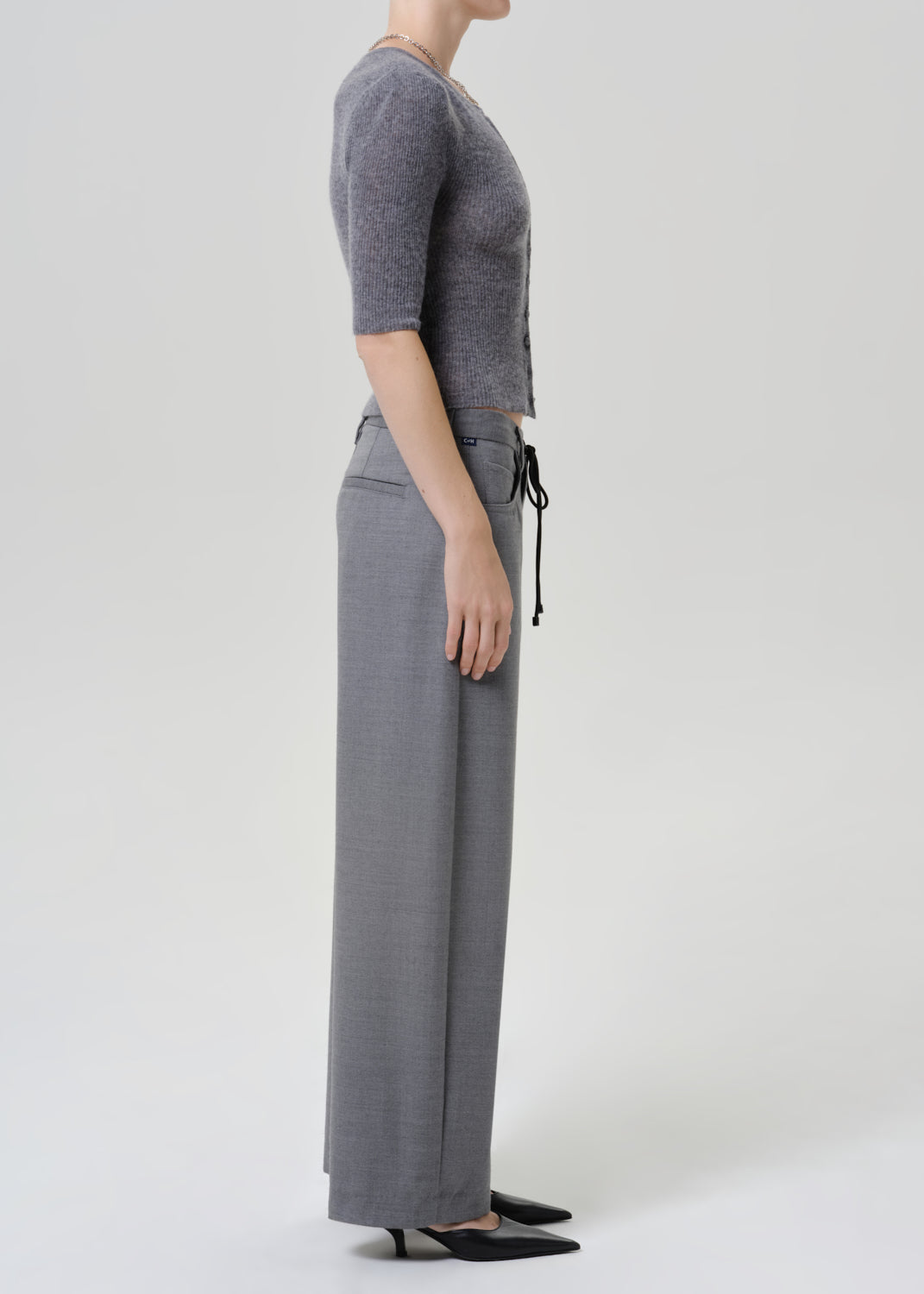 A model shown in profile wearing a gray short-sleeve knit top and matching gray wide-leg trousers with a drawstring waist, paired with black pointed flats, against a light gray studio background.