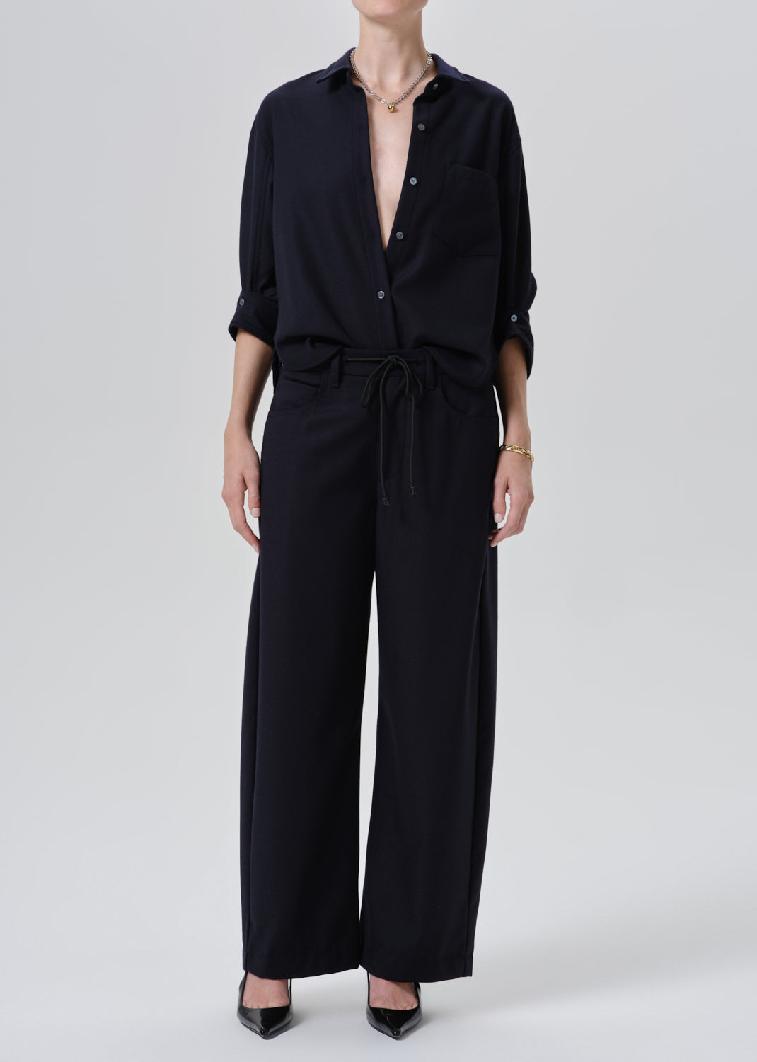 A model wearing a black wide-leg jumpsuit with a button-down shirt collar, rolled sleeves, and a drawstring waist, paired with black pointed-toe heels against a grey background.