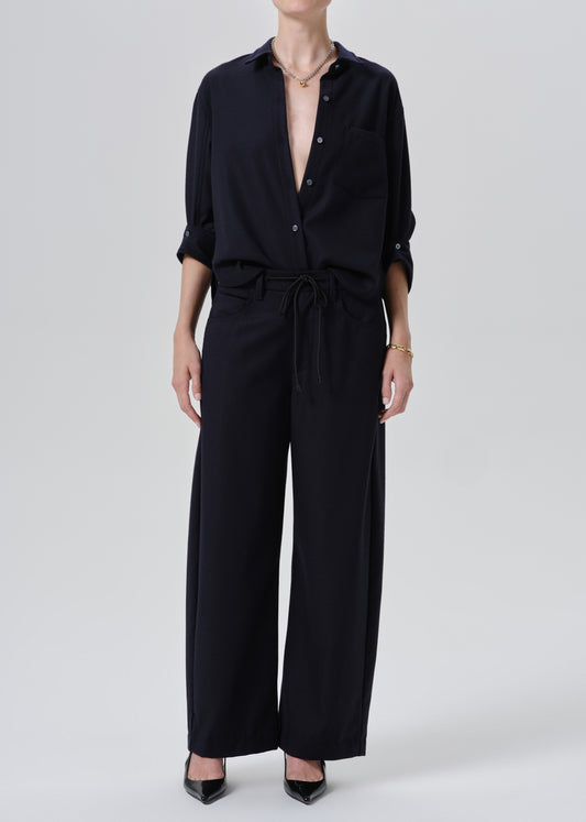 A model wearing a black wide-leg jumpsuit with a button-down shirt collar, rolled sleeves, and a drawstring waist, paired with black pointed-toe heels against a grey background.