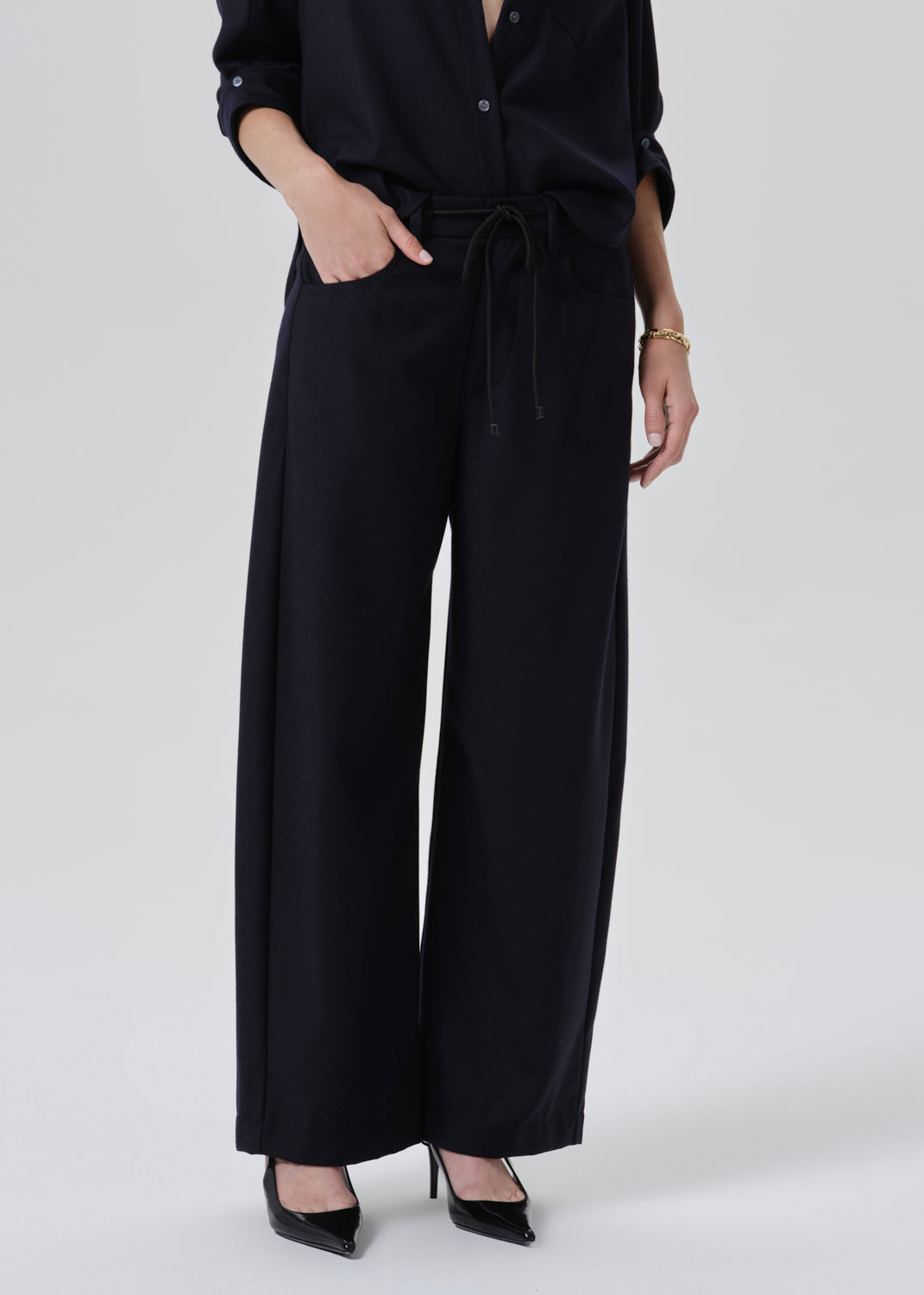 A model wearing black wide-leg trousers with a drawstring waist, paired with a black long-sleeve top and black pointed-toe heels, photographed against a grey background.