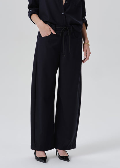 A model wearing black wide-leg trousers with a drawstring waist, paired with a black long-sleeve top and black pointed-toe heels, photographed against a grey background.