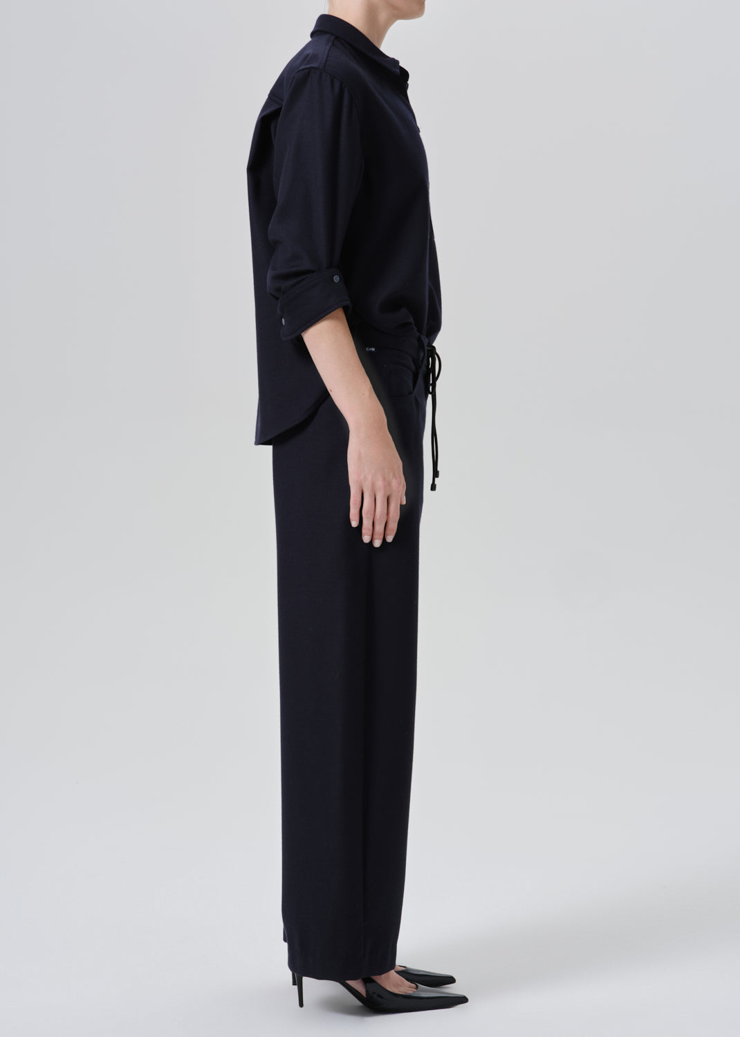 A model shown from the side wearing a dark navy blue shirt and matching wide-leg trousers with a drawstring waist, paired with black pointed-toe heels, against a grey background.