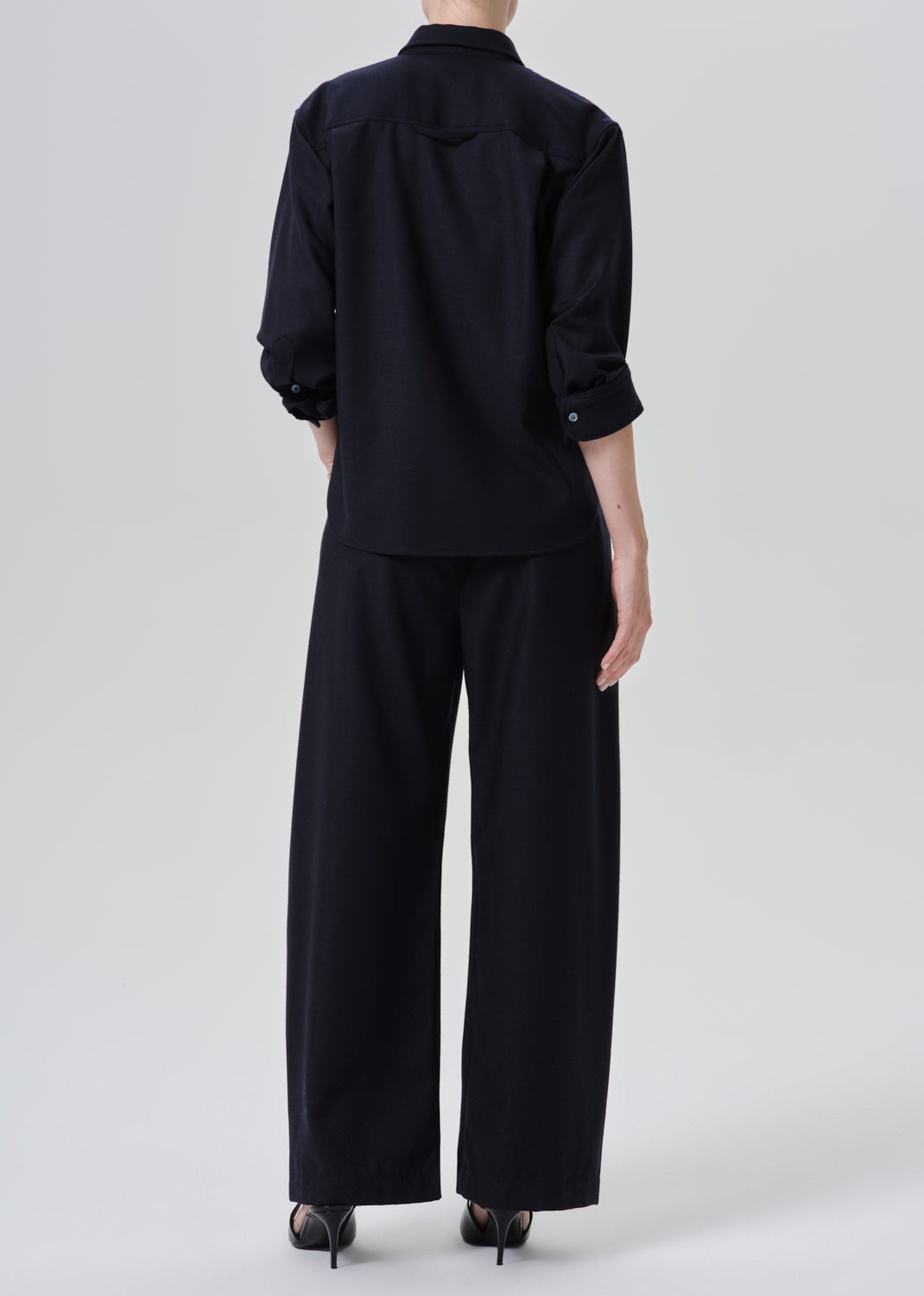 A model shown from the back wearing a dark navy blue shirt and wide-leg trousers set, paired with black heels, against a grey background.