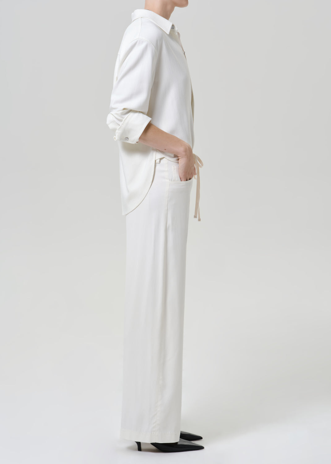 Woman wearing white wide leg pants with a white button-up shirt viewed from the side on a grey background.