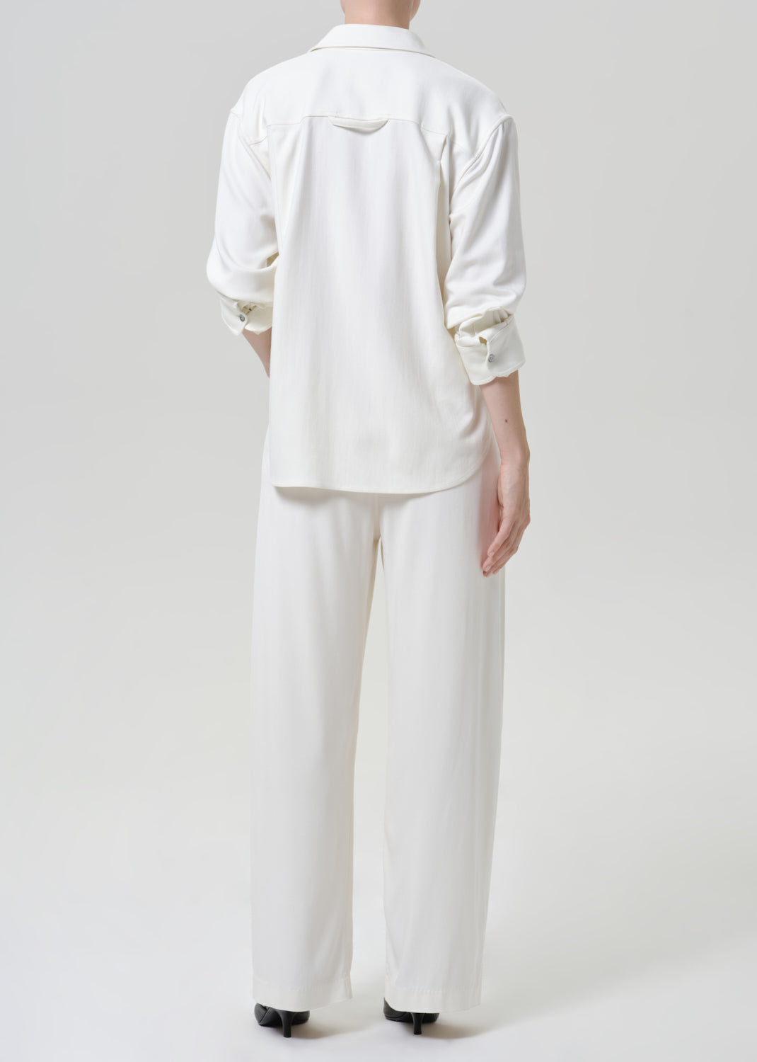 Woman wearing white wide leg pants with a white button-up shirt viewed from the back on a grey background.