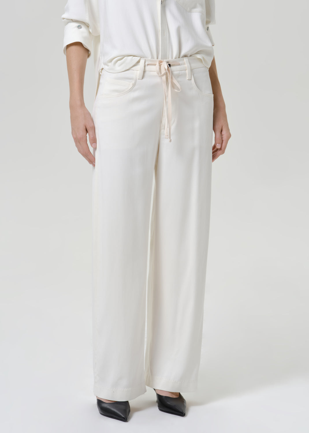 Woman wearing white wide leg pants with a white button-up shirt on a grey background.