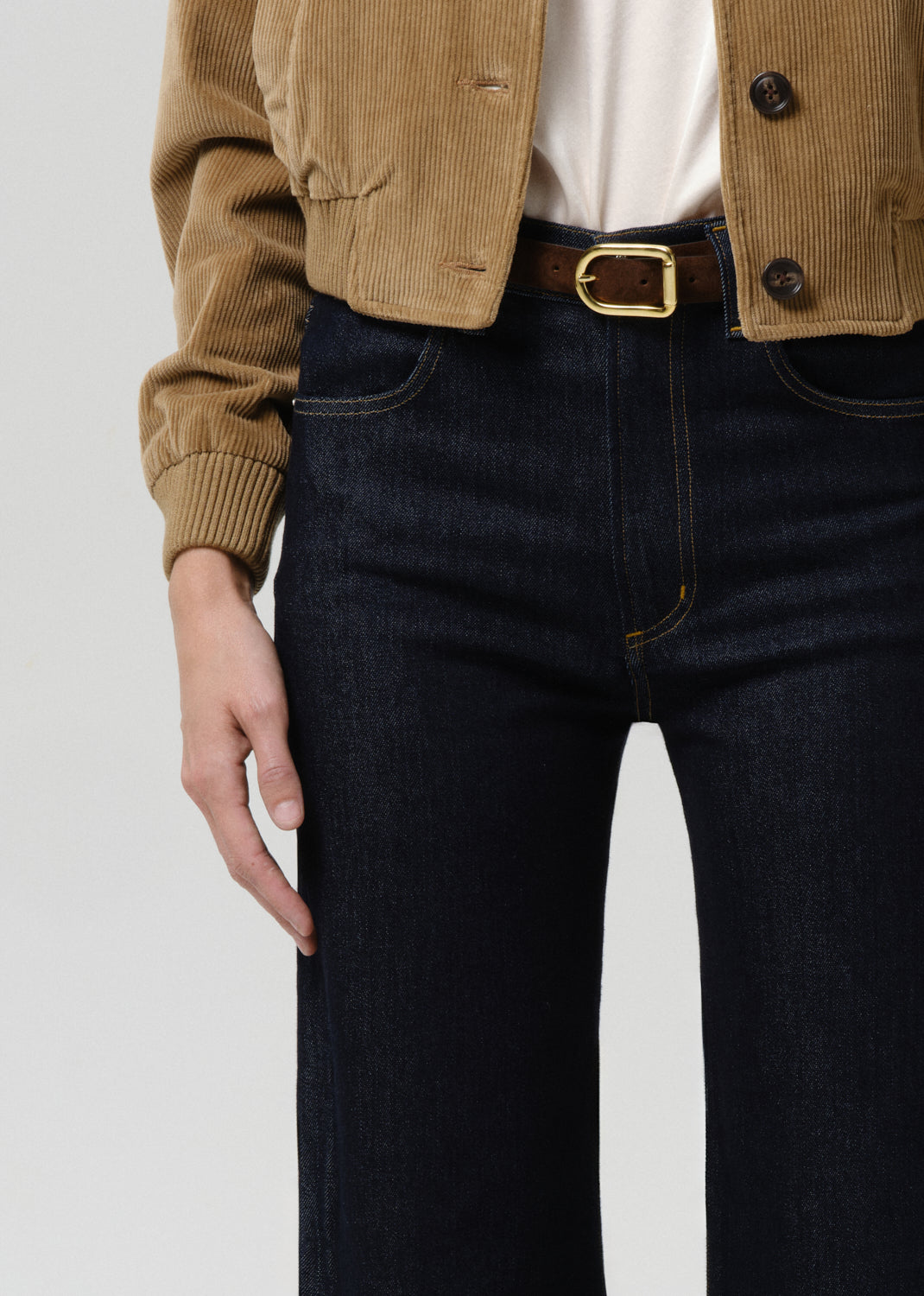 Person wearing a brown jacket and dark jeans with a gold belt buckle on a grey background