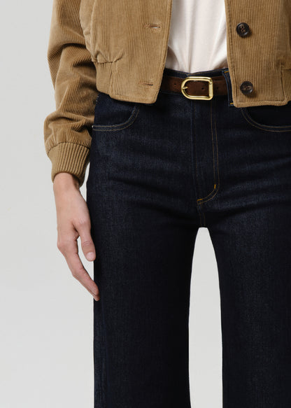 Person wearing a brown jacket and dark jeans with a gold belt buckle on a grey background