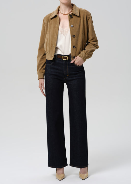 Person wearing a brown jacket, white top, black pants, and beige shoes on a grey background