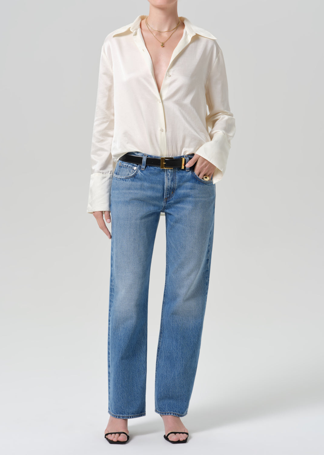 A model wearing a cream-colored satin button-down shirt and light blue straight-leg jeans with a black belt, standing against a grey background.