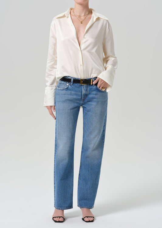 A model wearing a cream-colored satin button-down shirt and light blue straight-leg jeans with a black belt, standing against a grey background.