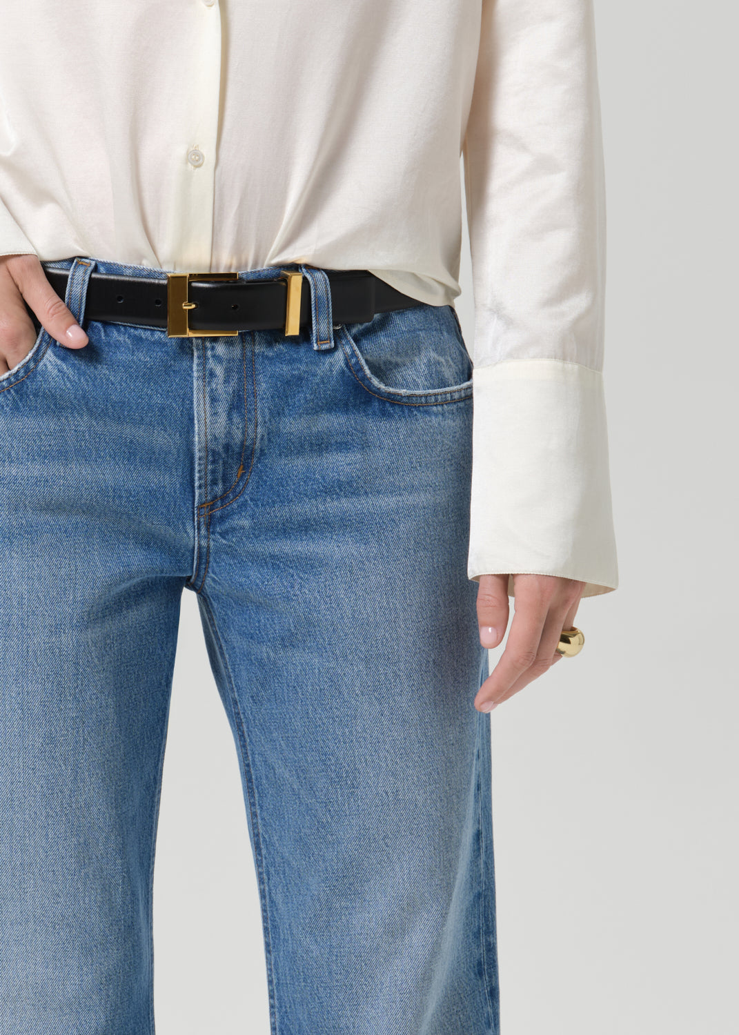 A close-up of a person wearing blue jeans with a black leather belt featuring a gold-tone rectangular buckle, paired with a cream-colored long-sleeve blouse.