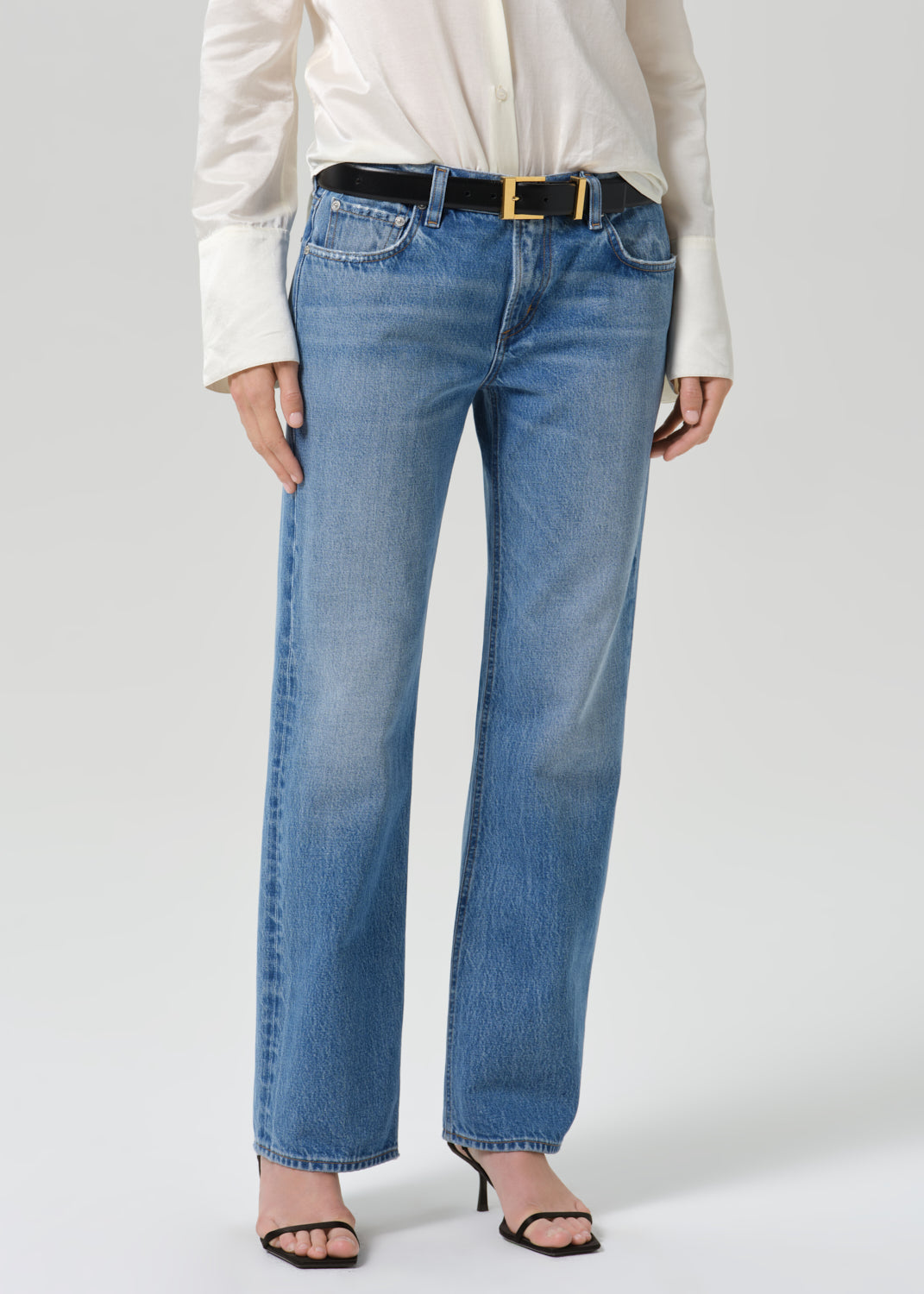 A model wearing a pair of light blue straight-leg denim jeans with a black belt featuring a gold buckle, styled with a white blouse and black heeled sandals against a grey background.