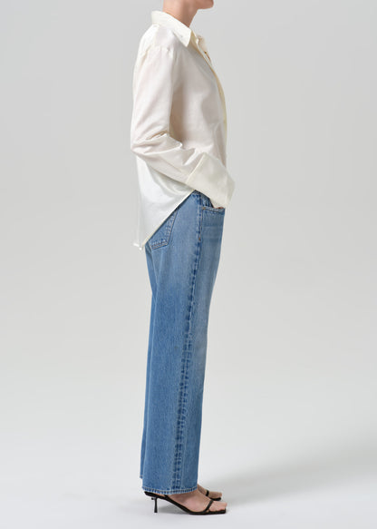 A side-profile view of a model wearing a white button-up shirt and light blue wide-leg jeans, paired with black strappy heels, against a grey background.