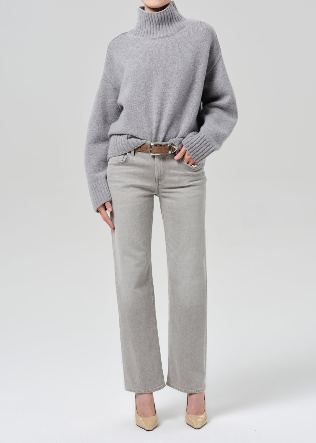 Person wearing a light grey oversized turtleneck sweater with light grey straight leg jeans on a grey background