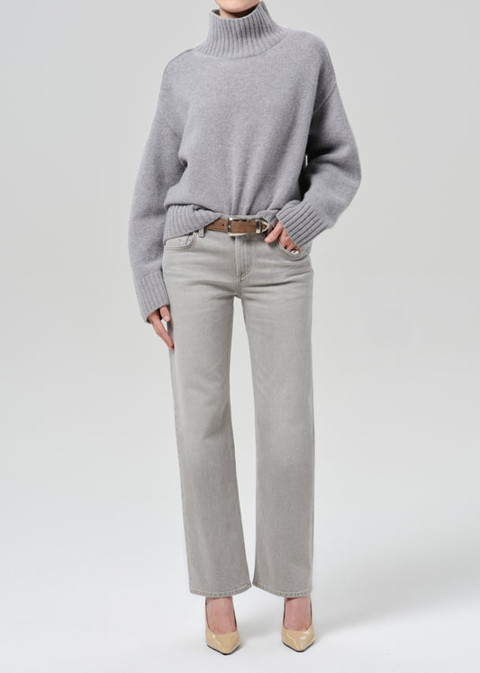 Person wearing a light grey oversized turtleneck sweater with light grey straight leg jeans on a grey background