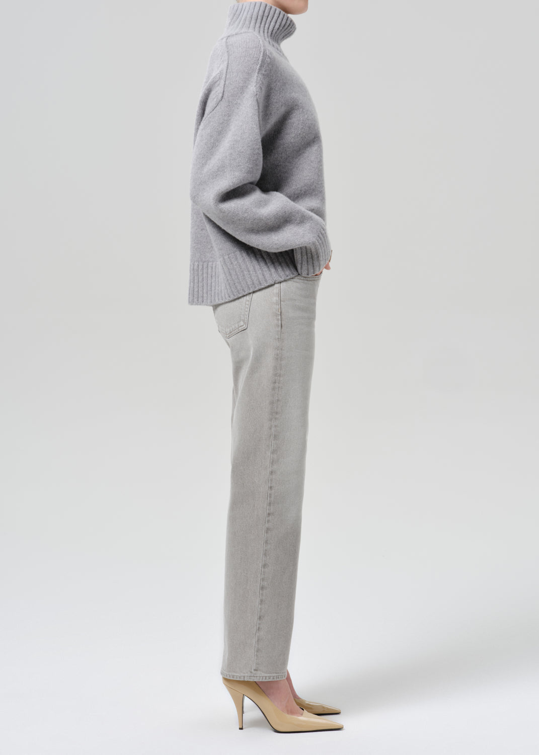 Person wearing a light grey oversized turtleneck sweater with light grey straight leg jeans viewed from the side on a grey background