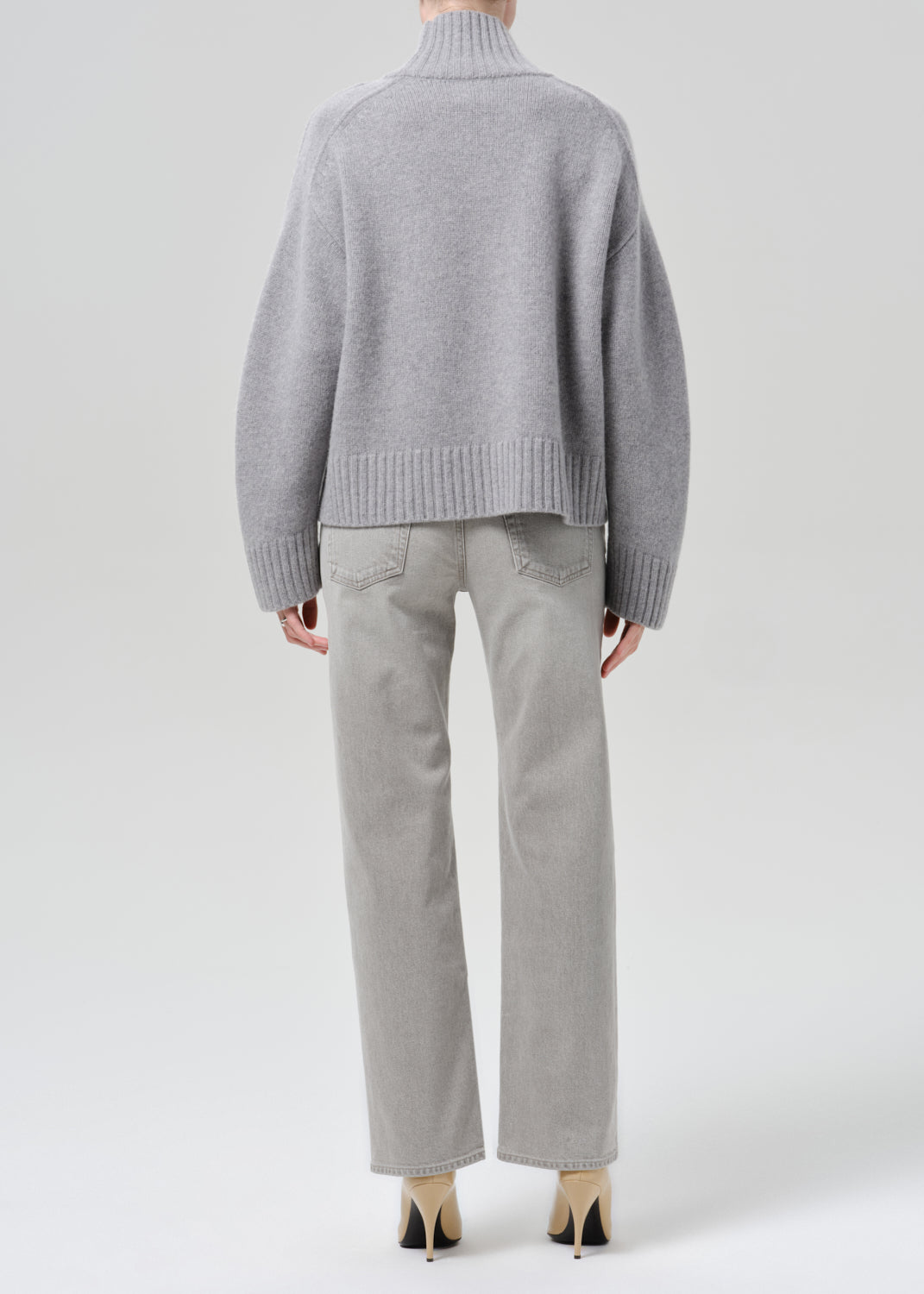 Person wearing a light grey oversized turtleneck sweater with light grey straight leg jeans viewed from the back on a grey background
