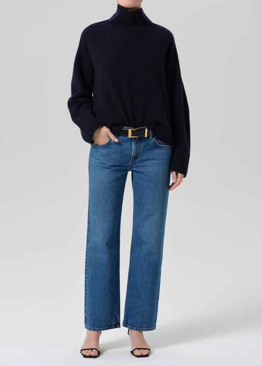 A model wearing a dark navy blue turtleneck sweater, blue wide-leg jeans with a gold belt, and black sandals, standing against a grey background.
