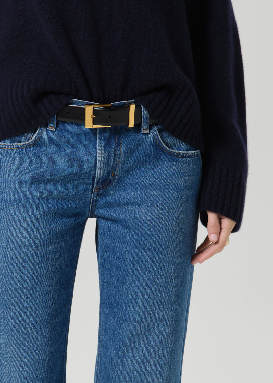 Close-up of a person wearing a black belt with a gold rectangular buckle, styled with blue jeans and a dark navy sweater against a grey background.