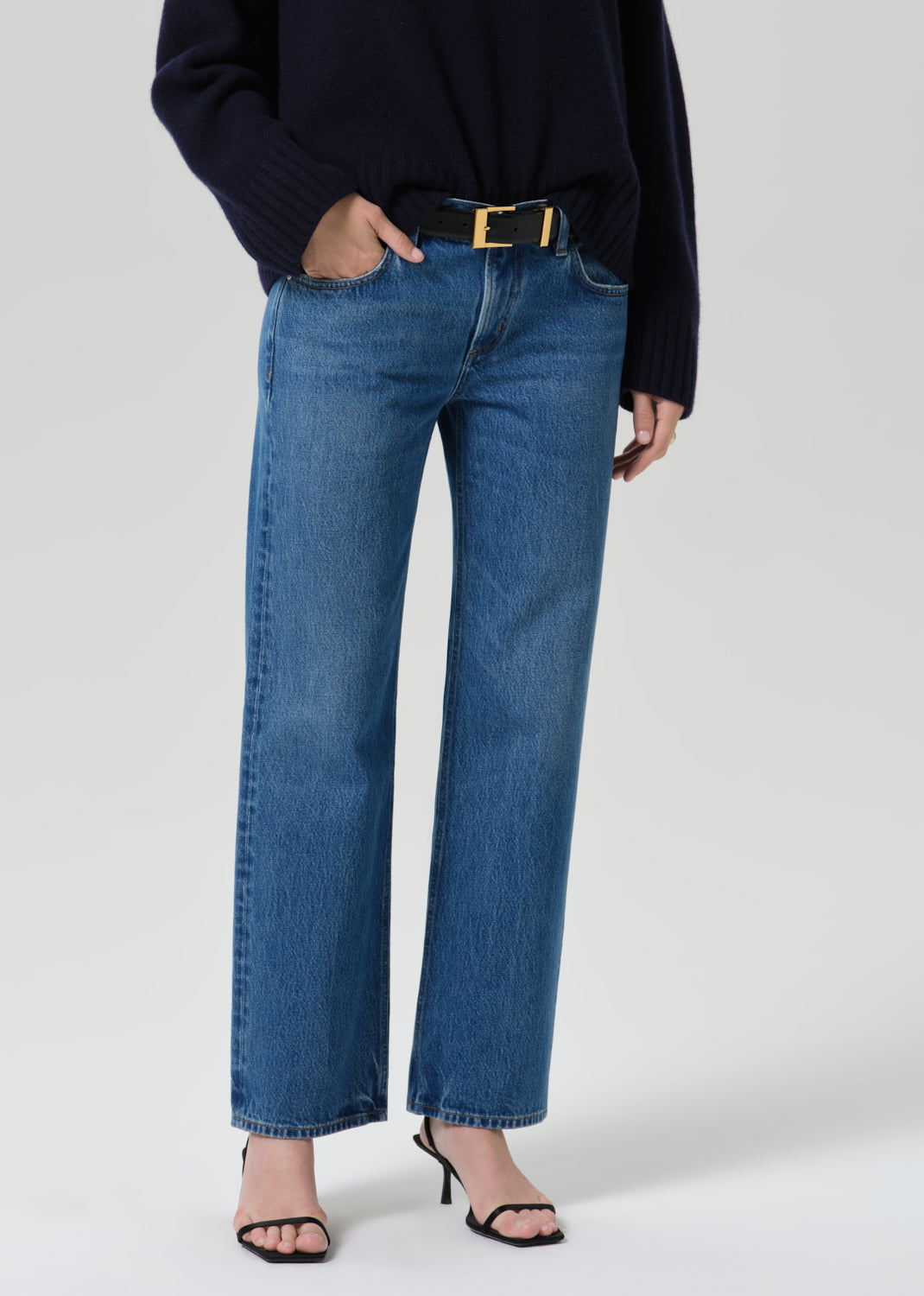 A model wearing a pair of medium-wash blue straight-leg jeans with a gold-tone belt, styled with a dark navy sweater and black strappy heels against a grey background.